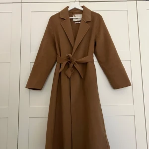 Handgjord kappa från MNG med bälte - Snygg, handgjord kappa från Mango som endast använts ett fåtal gånger. Kappan är tillverkad i ullblandning och har en stilren, rak passform med långa ärmar. Nypris 1 799kr