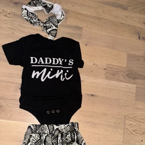 Svart body Daddy's Mini med ormmönstrade shorts - Svart kortärmad body med vit text 'Daddy's Mini' framtill. Tryckknappar i grenen för enkel påklädning. Tillhörande shorts och hårband i beige och svart ormmönster. Perfekt set för en cool och trendig look till de minsta.🥰