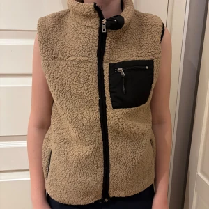 Beige fleeceväst från 157 - Mysig beige fleeceväst från 157 med svart bröstficka och dragkedja framtill. Västen har hög krage och svarta detaljer vid fickor och kanter. Perfekt för lager-på-lager och chill dagar.
