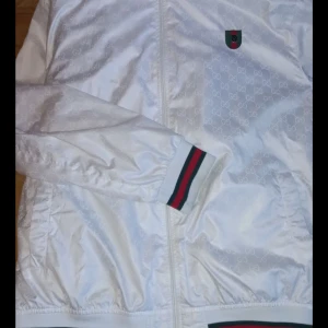 Vit vindjacka från Gucci äkta - Snygg vit vindjacka från Gucci med diskret GG-mönster i tyget och klassiska gröna och röda ränder på muddar och midja. Jackan har dragkedja framtill och en liten broderad logga på bröstet. Perfekt för dig som vill sticka ut med en lyxig streetstyle.