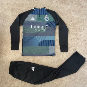 Real Madrid långärmad träningströja Adidas - Säljer en långärmad Real Madrid träningströja från Adidas med patchworkmönster i blått, grönt och svart. Tröjan har dragkedja vid halsen, klubbmärke och Adidas-logga på bröstet samt trycket 'Emirates Fly Better'. Tillverkad i funktionsmaterial.