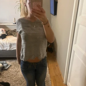 Grå stickad croppad topp - Säljer en grå stickad topp med kort ärm och croppad passform. Toppen har vågig nederkant och ett diskret randigt mönster i stickningen. Perfekt till jeans eller kjol för en chill look. Tröjan har ingen lapp så vet inte storlek men chansar på s eftersom jag brukar ha det.
