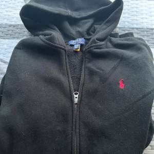 Polo Ralph Lauren hoodie - Svart hoodie från Polo Ralph Lauren med dragkedja och huva. Klassisk röd logga broderad på bröstet och stora fickor framtill. Mjuk bomullskvalitet, perfekt för chill dagar eller när du vill ha en clean streetstyle-look.