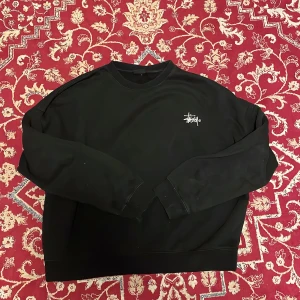 Oversized Stüssy sweatshirt - Svart sweatshirt från Stüssy med klassisk vit logga på bröstet och en stor vit Stüssy-grafik på ryggen. Tröjan har rund hals, långa ärmar och är gjord i mjuk bomullsmix. Perfekt för en streetwear-look.