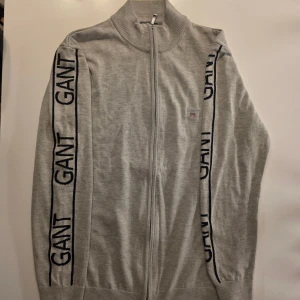 Grå kofta med dragkedja från GANT - Snygg grå kofta från GANT i 100% bomull med hel dragkedja och ribbade muddar. Svarta GANT-loggor längs båda ärmarna och liten broderad logga på bröstet. Perfekt för lager-på-lager och chill stil.           Väldigt snygg och gör dig bredare i axlarna sitter perfekt som har storlek M fast den är slim fit L