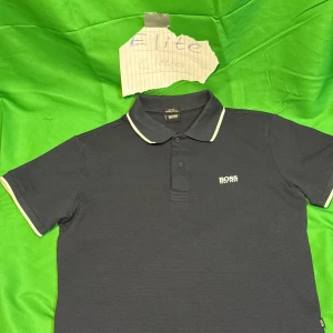 Svart pikétröja  BOSS - Snygg svart pikétröja BOSS med vit logga på bröstet och vita detaljer på krage och ärmslut. Klassisk pikékrage och korta ärmar, perfekt för en clean och stilren look. Tillverkad i bomull för skön känsla.Liknande modell som originalet!