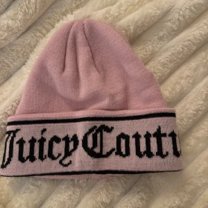 Rosa mössa från Juicy Couture - Snygg rosa mössa från Juicy Couture med bred uppvikt kant och svart logotyp i gotisk stil. Tillverkad i mjuk stickad bomullsmix, perfekt för att hålla värmen och ge en trendig touch till din outfit.