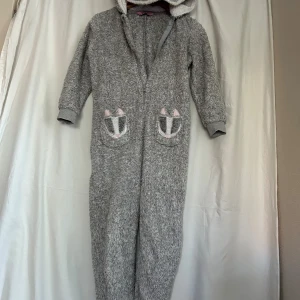 Grå fluffig onesie från Hunkemöller - Supermysig grå onesie från Hunkemöller med huva som har broderat kattansikte och öron. Fickor framtill med söta detaljer och dragkedja. Perfekt för chilliga dagar hemma eller sleepovers.