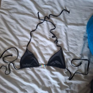 Svart bikiniöverdel med pärldetalj - Svart trekantsbikini med knytband i nacken och ryggen. Mellan kuporna sitter färgglada pärlor som ger en söt touch. Klassisk modell som är enkel att justera till rätt passform. Aldrig använd men har tagit bort lappen.