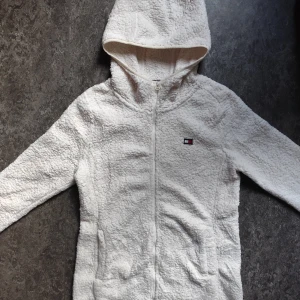 Tommy Hilfiger Fleece Huvtröja - Mysig vit Fleece huvtröja från Tommy Hilfiger med huva och dragkedja framtill. Huvtröjan har två fickor och ett litet broderat logomärke på bröstet. Perfekt för kyliga dagar och riktigt skön att ha på sig. Snygga detaljer med Hilfigers klassiska färger på insidan av kragen.