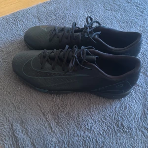 Svarta Nike fotbollsskor med snörning - Säljer ett par svarta Nike fotbollsskor med klassisk snörning och låg profil. Skorna har en diskret Nike-logga på sidan och mönstrad sula för bra grepp på planen. Tillverkade i syntetmaterial med en stilren look, perfekta för dig som vill ha snygga och funktionella skor till fotbollen. Knappt använd 