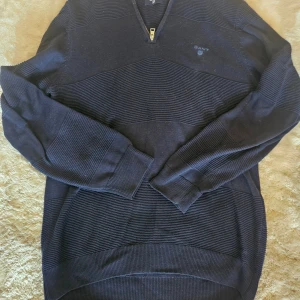 Mörkblå stickad tröja från GANT - Snygg mörkblå stickad tröja från GANT med diskreta horisontella ränder och half zip-dragkedja vid halsen. Tröjan har lång ärm och klassisk passform, perfekt för dig som gillar stilrena och tidlösa plagg.