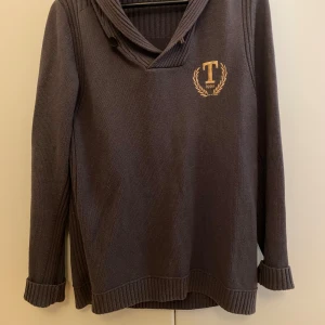 Mörkgrå stickad tröja med emblem - Mysig mörkbrun stickad tröja med snyggt broderat emblem på bröstet. Tröjan har en unik krage med knappdetalj och långa ärmar med ribbade muddar. Perfekt för kyliga dagar och en stilren look.