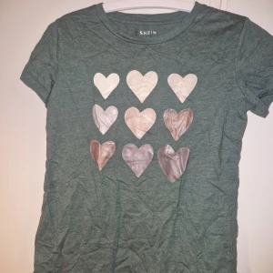 Grön t-shirt med hjärtan  - Grön t-shirt från SHEIN med korta ärmar och rund hals. Framsidan är dekorerad med nio hjärtan i olika nyanser av vitt, beige och grått. Tshirten har en avslappnad passform, perfekt för en chill vardagslook.