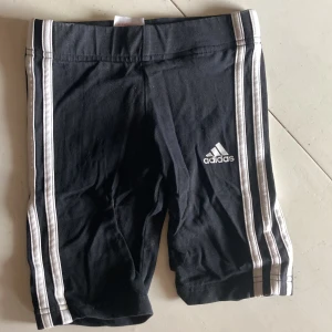 Svarta Adidas shorts med vita ränder - Svarta shorts från Adidas med klassiska vita ränder längs sidorna och Adidas-logga framtill. Modellen är tight och passar perfekt till träning eller sport. Tillverkade i mjuk bomull för maximal komfort.
