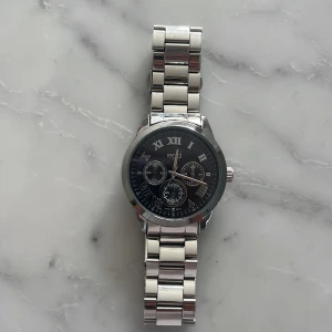 Regal silverfärgad klocka med länk - Stilren klocka från Regal med silverfärgad länk i metall och svart urtavla. Urtavlan har romerska siffror och tre mindre dekorativa subdials. Klassisk design som passar till många olika stilar. Metallarmbandet har en smidig låsmekanism.