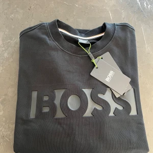 Svart sweatshirt från BOSS - Svart sweatshirt från BOSS med stort, upphöjt logotryck på bröstet. Tröjan har rund halsringning, långa ärmar och ribbade muddar. Tillverkad i mjuk bomullsmix som känns skön mot huden. Perfekt för en clean och stilren look.