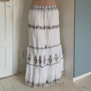Vit långkjol med broderi från Zara - Supersnygg vit långkjol från Zara med broderade detaljer i svart och beige. Kjolen har resår och snörning i midjan, samt flera broderade band och mönster längs kjolen. Kjolen är 94 cm lång och midjemåttet mätt rakt över är 40 cm. Dock är det resor upptill, alltså passar den om man har ännu bredare höfter! 