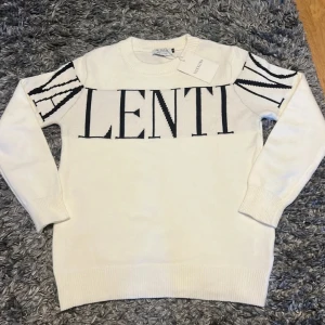 Vit stickad tröja från Valentino - Vit stickad tröja från Valentino med svart logotyp över bröstet och axlarna. Tröjan har rund halsringning, ribbade muddar och lång ärm. Snygg och stilren design som sticker ut med den stora texten.