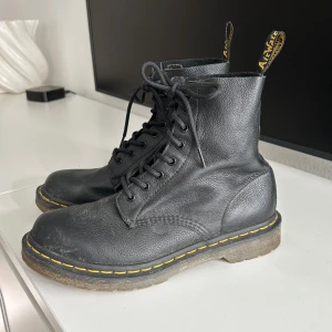 Svarta Dr. Martens boots i skinn - Klassiska svarta Dr. Martens boots i grovt skinn med snörning och ikonisk gul söm runt sulan. Robust platt sula och dragflik baktill med logga. Defekter på skosnöret (sista bilden)