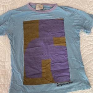 Blå t-shirt Les Coyotes de Paris - Snygg ljusblå t-shirt från Les Coyotes de Paris med lila ribbad krage och stor grafisk print i lila och brunt framtill. T-shirten har korta ärmar och är gjord i mjuk bomull. Perfekt för dig som gillar unika prints och coola färgkombinationer.