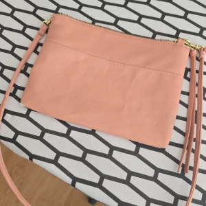 Ljusrosa axelväska från H&M - Söt ljusrosa axelväska från H&M med guldiga metalldetaljer och dragkedja upptill. Väskan har en slät ovandel och en lätt strukturerad nederdel, samt en lång justerbar rem. Några skav märken se bilder men ändå inte mkt fräscht skick. Tror de uppkommit av trångt utrymme i garderoben 