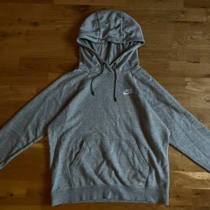 Grå Nike hoodie med broderad logo - Snygg grå hoodie från Nike med broderad vit logo på bröstet. Klassisk känguruficka framtill, justerbar huva med snören och mjukt material i bomullsblandning. Perfekt för chill eller streetwear. Långärmad och normal passform.
