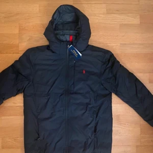 Svart pufferjacka från Polo Ralph Lauren - Svart pufferjacka från Polo Ralph Lauren med röd broderad logga på bröstet. Jackan har huva, dragkedja framtill och två sidofickor. Materialet är syntet och jackan är långärmad med en clean och modern look.