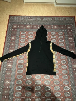 Svart Burberry hoodie med rutiga detaljer - Svart hoodie från Burberry med klassiska beige-rutiga paneler längs sidorna och ärmarna. Tröjan har huva med snörning, känguruficka framtill och långa ärmar. Perfekt för dig som vill ha en snygg streetwear-look med lyxig touch.