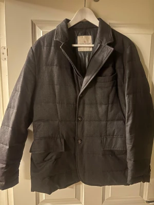 Moncler jacka - Snygg mörkgrå dunjacka från Moncler med quiltad design och klassisk kavajkrage. Jackan har två stora fickor framtill, bröstficka och stängs med knappar samt dragkedja. Materialet är polyester och dun, vilket gör den både varm och stilren för kalla dagar. Storlek 3