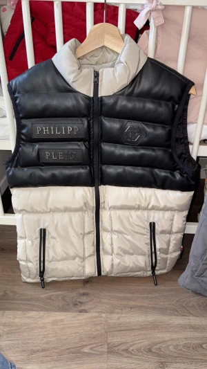 Svart och beige dunväst Philipp Plein - Snygg dunväst från Philipp Plein i svart och beige med quiltad design. Västen har hög krage, dragkedja framtill och två fickor med dragkedja. Märkeslogga och broderade detaljer på bröstet. Materialmix av skinn och polyester för en lyxig känsla. Skriv privat för mer information!