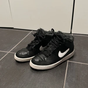Svarta Nike sneakers med fuskpäls - Svarta Nike sneakers med vit swoosh och vit sula. Skorna har snörning och är fodrade med mjuk, mörkgrå fuskpäls runt hela öppningen för extra värme. Ovandelen är i skinn och de har en klassisk rund tå. Perfekta för dig som vill ha streetstyle med cozy vibe. Fungerar som barnsko, storlek 36.
