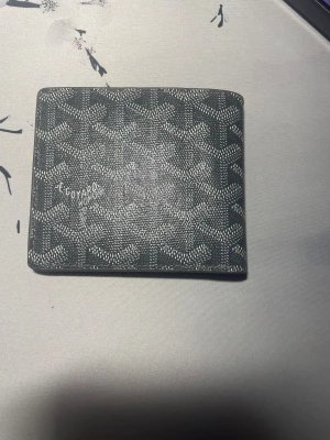 Svart Goyard plånbok med mönster - Snygg svart plånbok från Goyard med deras ikoniska grafiska mönster i vitt. Plånboken är stilren och har en klassisk fyrkantig form, perfekt för kort och sedlar. Materialet är skinn och designen är riktigt clean och modern.