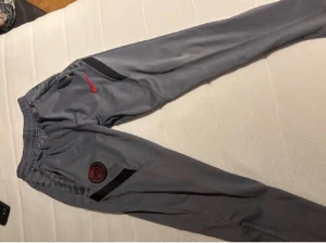 Grå Nike PSG mjukisbyxor - Snygga grå mjukisbyxor från Nike med PSG-logga på benet och röd swoosh. Byxorna har svarta detaljer längs sidorna och elastisk midja för skön passform. Perfekta för träning eller chill. Slimmad modell med sportig vibe.