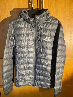 Grå pufferjacka EA7 Emporio Armani - Snygg grå pufferjacka från EA7 Emporio Armani med huva och tydlig logga på bröstet. Jackan har quiltad design, dragkedja framtill med EA7-detalj och är tillverkad i syntetmaterial. Perfekt för dig som gillar stilren och sportig look. Liten i storlek. Bredd=52, längd=66 och ärmlängd=48 cm.