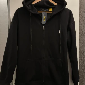 Polo Ralph Lauren zip up hoodie  - Svart hoodie från Polo Ralph Lauren med dragkedja framtill, huva med snörning och klassisk broderad logga på bröstet. Tillverkad i mjuk bomull och har två fickor framtill. Perfekt för en chill och stilren look.