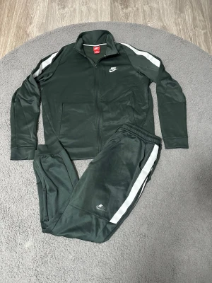 Nike träningsset i mörkgrönt, stl L - Nike träningsset i mörkgrönt med vita ränder längs ärmar och ben. Jackan har dragkedja och ståkrage, byxorna har resår i midjan. Båda delarna har Nike-logga tryckt i vitt. Perfekt för dig som gillar sportig och clean stil.