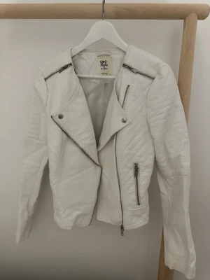 Vit skinnjacka från Vero Moda - Snygg vit skinnjacka från Vero Moda med quiltade detaljer på axlar och ärmar. Jackan har asymmetrisk dragkedja, silverfärgade blixtlås och knappar samt två fickor med dragkedja. 