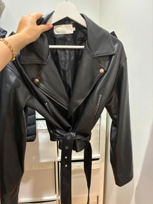 Svart skinnjacka med bälte från NLY Trend - Snygg svart skinnjacka från NLY Trend i bikerstil med breda slag, silverfärgade dragkedjor och knappar. Jackan har ett matchande bälte i midjan och långa ärmar. Perfekt för dig som vill ha en cool och edgy look.