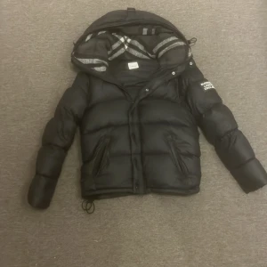 Svart pufferjacka från Burberry - Svart pufferjacka från Burberry med huva och klassiskt rutigt foder. Storlek S/XS. Jackan har dragkedja och knappar framtill, snörning nedtill och två sidofickor. På ena ärmen finns Burberry London England-tryck. Perfekt för kalla dagar. Jackan går även att göra till en väst. Pris kan diskuteras!
