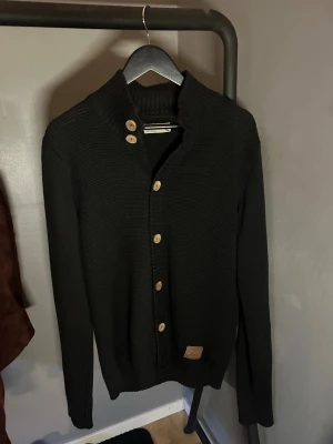 Svart stickad kofta från Solid - Svart stickad kofta från Solid med ribbad krage och träknappar längs hela framsidan. Koftan har långärmade ärmar och en liten patch vid nederkanten. Perfekt för lager-på-lager och chill stil.