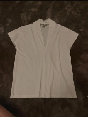 Vit v-ringad blus med korta ärmar - Säljer en stilren vit blus med v-ringad hals och korta ärmar. Toppen har en rak och loose passform, vilket gör den enkel att matcha med olika outfits. Materialet känns mjukt och lätt, perfekt för varmare dagar. Enkel och clean design utan mönster eller extra detaljer.