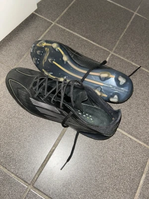 Svarta Adidas X fotbollsskor - Säljer ett par svarta Adidas X fotbollsskor med snörning och platt sula med dobbar. Skorna har en sportig design med vita detaljer och är tillverkade i syntetmaterial för lätt känsla på planen. Perfekta för dig som vill ha snabbhet och kontroll.