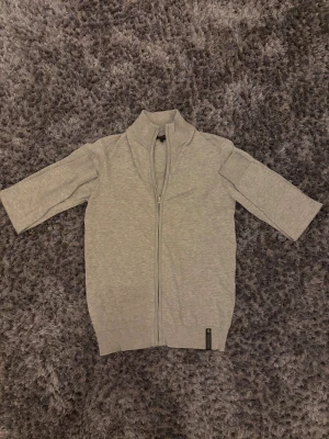 Grå kofta med dragkedja från Indicode - Snygg grå kofta från Indicode i storlek S. Koftan har en hög krage, trekvartsärm och hel dragkedja framtill. Tillverkad i mjuk bomull som känns skön mot huden. Perfekt för dig som gillar stilrena och enkla plagg.