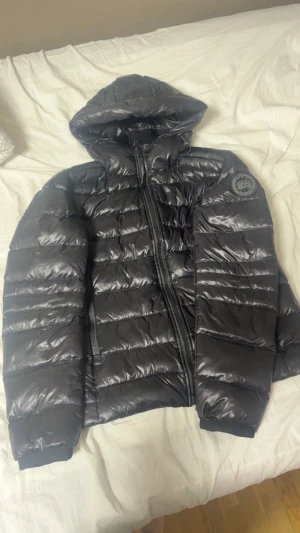 Canada Goose crofton black label - Säljer en svart pufferjacka från Canada Goose med huva och snyggt märke på ärmen. Jackan har glansig yta, dragkedja framtill med logotyp och quiltad design. Perfekt för kalla dagar och har en riktigt clean look.