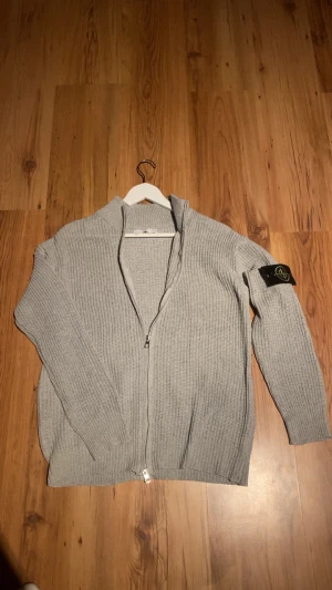 Grå stickad kofta från Stone Island - Säljer en grå stickad kofta från Stone Island med dragkedja framtill och ribbad struktur. Klassisk svart patch med logga på vänster ärm. Koftan har lång ärm och en clean, modern look som passar perfekt till jeans eller cargo. Fråga gärna om fler bilder eller info!