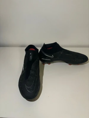 Nike Phantom GT svarta fotbollsskor - Nike Phantom GT fotbollsskor i svart med diskreta vita detaljer och röda dobbar. Skorna har en strumpliknande passform utan snörning, med syntetiskt material och texturerad yta för bättre bollkontroll. Snygg och modern design som sticker ut på planen. Pris kan diskuteras 