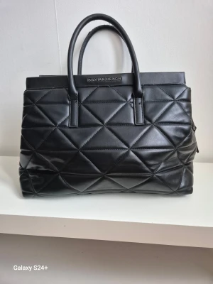 Svart quiltad handväska Silvian Heach - Stilren svart handväska från Silvian Heach med quiltad design och rejäla handtag. Väskan är tillverkad i skinn och har en praktisk dragkedja upptill. Insidan har fack med dragkedja för extra förvaring. Perfekt för dig som gillar klassisk och edgy look. OBS se bild på handtag... 