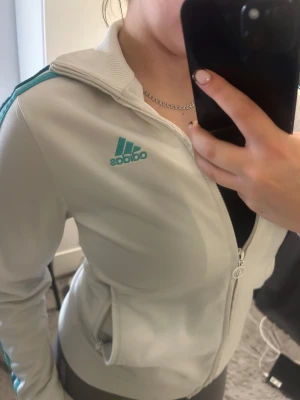 Vit Adidas vindjacka med turkosa ränder - Säljer en fräsch vit Adidas vindjacka med turkosa ränder längs ärmarna och Adidas-logga på bröstet. Jackan har ribbade muddar, hög krage och dragkedja framtill. Perfekt för dig som gillar sportig stil och vill sticka ut med klassiska detaljer.