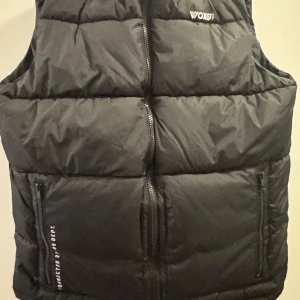 Svart dunväst från Woxon - Svart dunväst från Woxon med quiltad design och hög krage. Västen har två fickor med dragkedja och vit textdetalj på ena fickan. Perfekt för lager-på-lager och har en stilren look med diskret logga på bröstet.